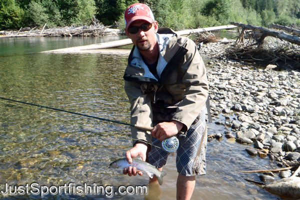 Photo of a fisherman by a river holding a small rainbow trout.